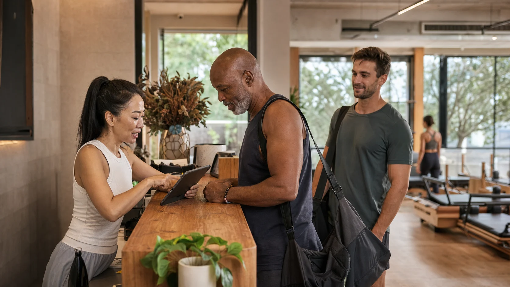 A studio manager helping an older member check in while another member waits at a bright Australian Pilates studio reception